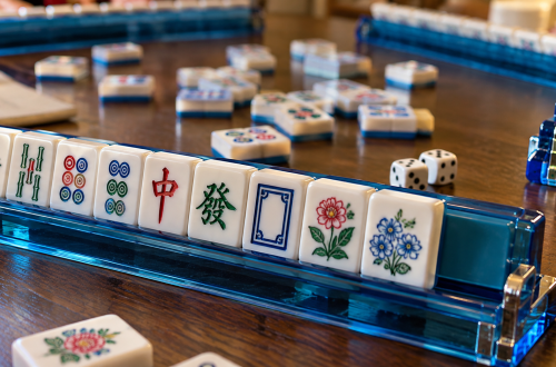 Close-up of American mah-jongg tiles arranged on a blue rack, with dice, discarded tiles, and other players’ racks visible on a wooden table.