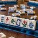Close-up of American mah-jongg tiles arranged on a blue rack, with dice, discarded tiles, and other players’ racks visible on a wooden table.